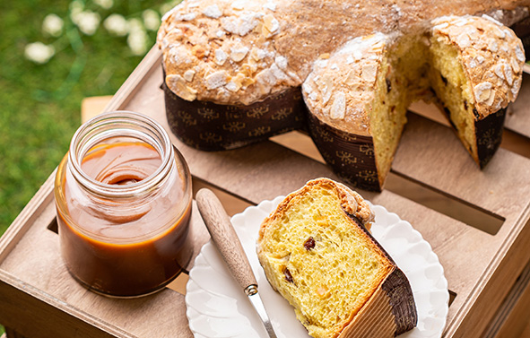Brioche colomba breads placed on a wooden crate, with a sliced piece on a plate, a jar of filling, and a knife beside it.