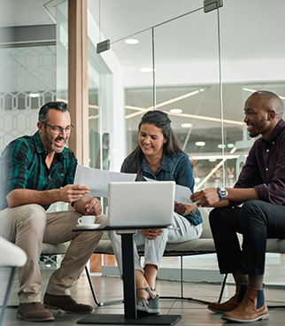 Trois personnes assises dans un espace de travail, regardant des documents et un ordinateur portable posé sur une table basse, dans un bureau vitré.