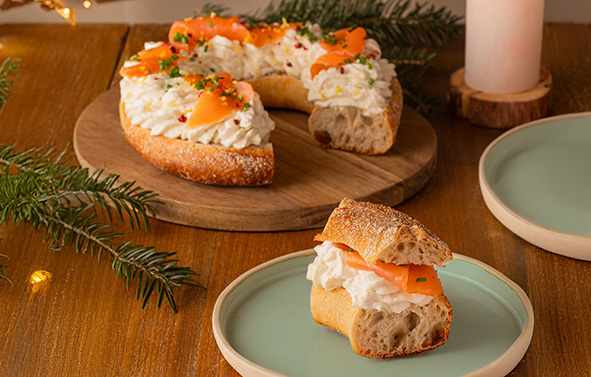 Pain en forme de couronne garni de fromage frais et de saumon, posé sur une planche en bois, avec une part servie sur une assiette à côté.