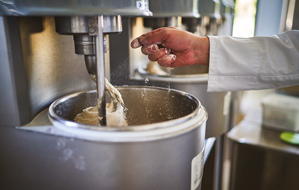Hand of a person adding an ingredient into a stainless-steel industrial mixer containing dough being mixed.