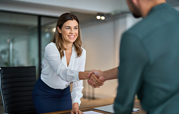 Femme souriante tendant la main pour serrer celle d’une autre personne, de l’autre côté d’un bureau, dans un espace de travail.