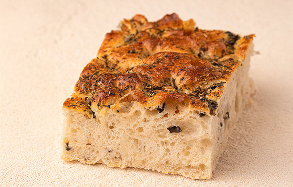 Piece of focaccia with an open crumb, featuring a golden crust topped with herbs, placed on a floured surface.