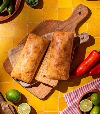 Two ciabatta-style breads placed on a wooden board, on a yellow tiled surface, surrounded by chili peppers and limes.