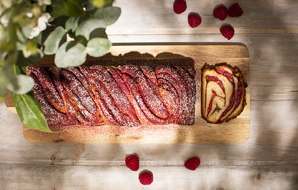 Babka marbrée à la framboise présentée sur une planche en bois, avec une tranche découpée et des framboises disposées autour.