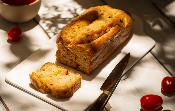 Rectangular savory tomato loaf cake, partially sliced, presented on a white dish with a knife beside it.