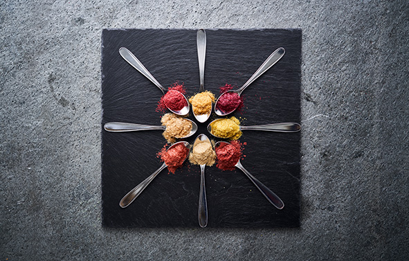 Spoons arranged in a circle on a black slate, containing powders of different colors, red and yellow, viewed from above.