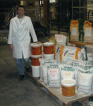 Hombre con bata blanca de pie en un taller industrial, junto a un palé cargado con sacos y cubos de productos.