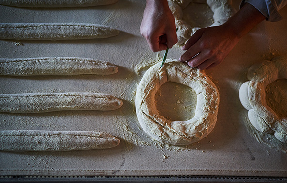 Mains en train de façonner une pâte farinée en forme de couronne sur un plan de travail, avec des pâtons allongés disposés à côté.