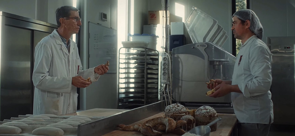Two professionals in laboratory attire taste and exchange around breads in a production workshop equipped with machines and baking trays.