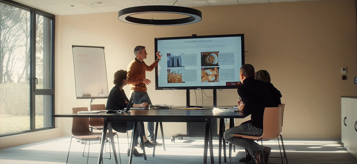 Reunión en una sala con un ponente presentando productos de panadería en una pantalla, frente a tres personas sentadas alrededor de una mesa.