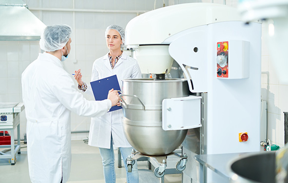 Deux personnes en blouse blanche et charlotte discutant devant un pétrin industriel en acier inoxydable, dans un atelier de production.