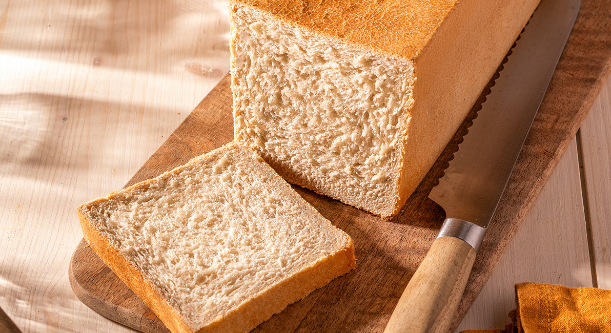 Rectangular sandwich loaf, partially sliced, placed on a wooden board with a knife beside it, showing a fine and even crumb.