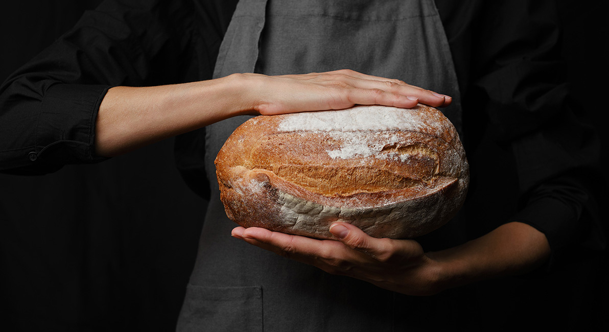 Person holding an oval country-style bread with a golden, lightly floured crust, against a dark background.