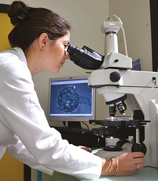 Persona con bata blanca observando una muestra con un microscopio en un laboratorio, con una pantalla de ordenador visible al fondo.