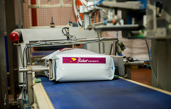 Bolsa de producto envasado sobre una cinta transportadora automática, en una línea de envasado industrial, con máquinas visibles alrededor.