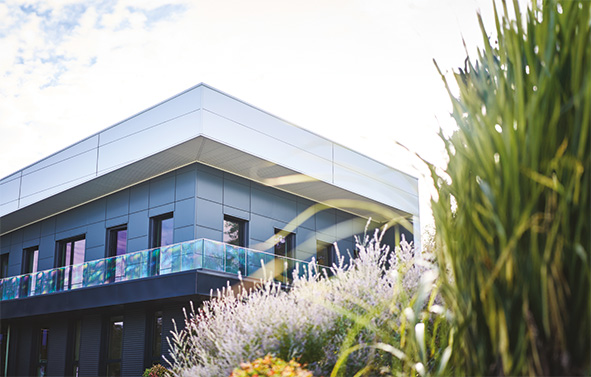 Bâtiment industriel moderne à deux niveaux, avec façade gris foncé et grandes fenêtres, entouré de plantes et de végétation au premier plan, sous un ciel clair.