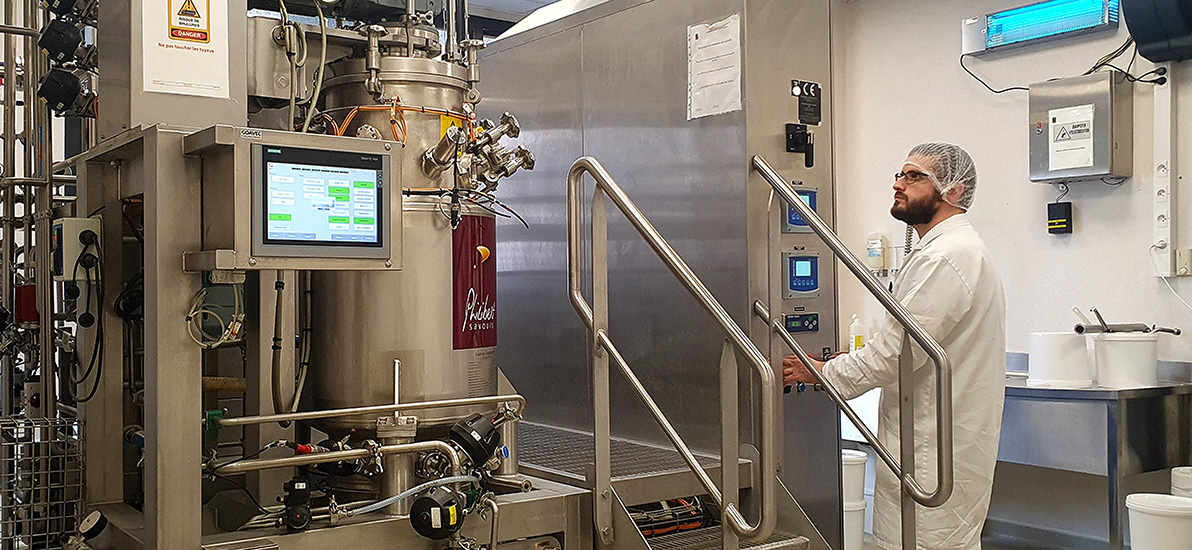 Person wearing a lab coat and hairnet operating an industrial fermentation machine with a control screen in a production laboratory.