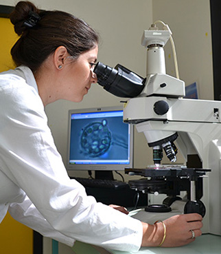 Opératrice en blouse de laboratoire observant un échantillon au microscope, dans un environnement de recherche équipé d’un écran d’analyse scientifique.