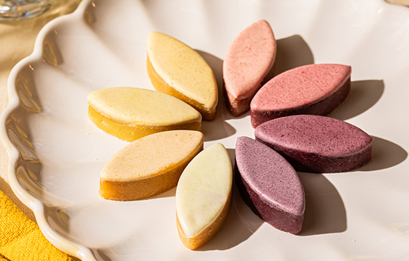 Assortiment de calissons colorés disposés en cercle sur une assiette blanche, aux teintes jaunes, orangés, roses et violettes.