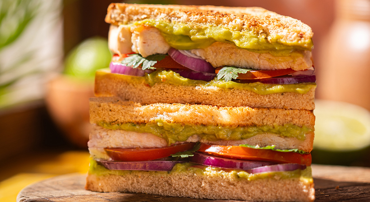 Sándwich compuesto de rebanadas de pan de molde rellenas de pollo, tomate, cebolla roja, hierbas y salsa verde, apilado sobre una tabla de madera.