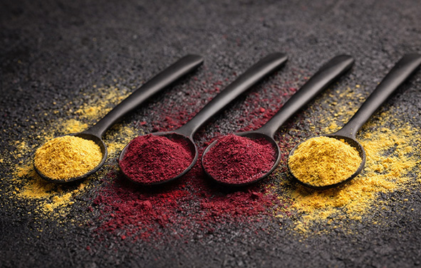 Four black spoons aligned, containing yellow and red powders, arranged on a dark surface with scattered powder around.